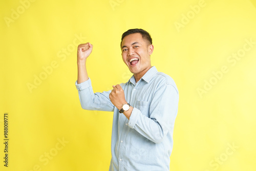 Excited Asian man in light blue denim shirt with fist pump hand isolated on yellow background