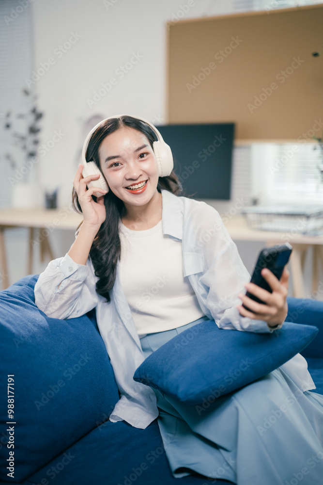 Young woman is relaxing on a comfortable blue sofa, enjoying her favorite music through wireless headphones while browsing social media on her smartphone Young woman is relaxing on a comfortable blue sofa, enjoying her favorite music through wireless headphones while browsing social media on her smartphone