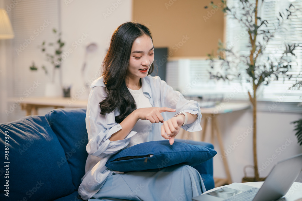 Obraz premium Young asian freelancer works from home, checking time on her smartwatch while using her laptop on the couch. She looks content and efficient in a modern work-from-home setup