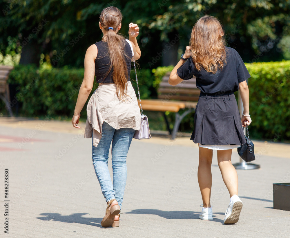 Fototapeta premium Two women walking down a sidewalk, one of them carrying a purse