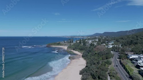 Aerial ascend of Coledale's sandy beaches and coastal road, surrounded by greenery and ocean, under a clear blue sky photo