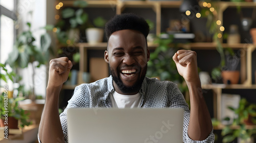 Happy young man in front of laptop computer raising hands and laughing excited about success. Emotional guy feeling glad and euphoric about getting work done and finishing successfu photo