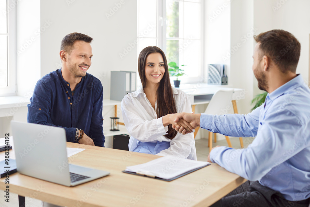 Wife and husband, as clients, shake hands with a consultant or agent, confirming a contract or deal during a meeting. Their handshake symbolizes the finalization of the agreement, contract or deal. Wife and husband, as clients, shake hands with a consultant or agent, confirming a contract or deal during a meeting. Their handshake symbolizes the finalization of the agreement, contract or deal.