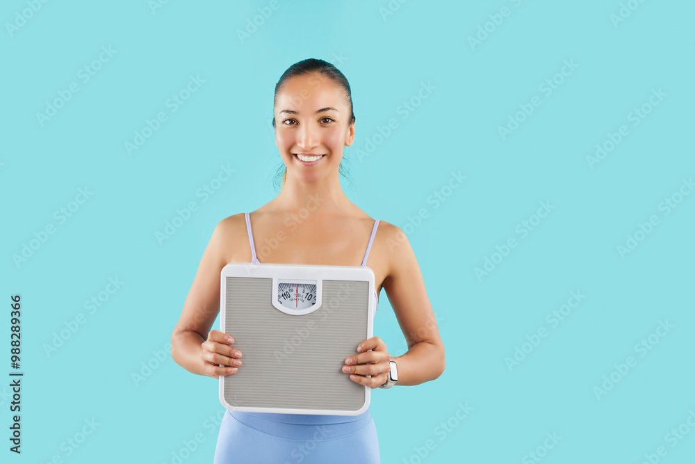 Woman holding scales and smiles, representing weight loss during sport or fitness workout and a healthy lifestyle. Her fitness and diet journey is highlighted on a blue background . Woman holding scales and smiles, representing weight loss during sport or fitness workout and a healthy lifestyle. Her fitness and diet journey is highlighted on a blue background .