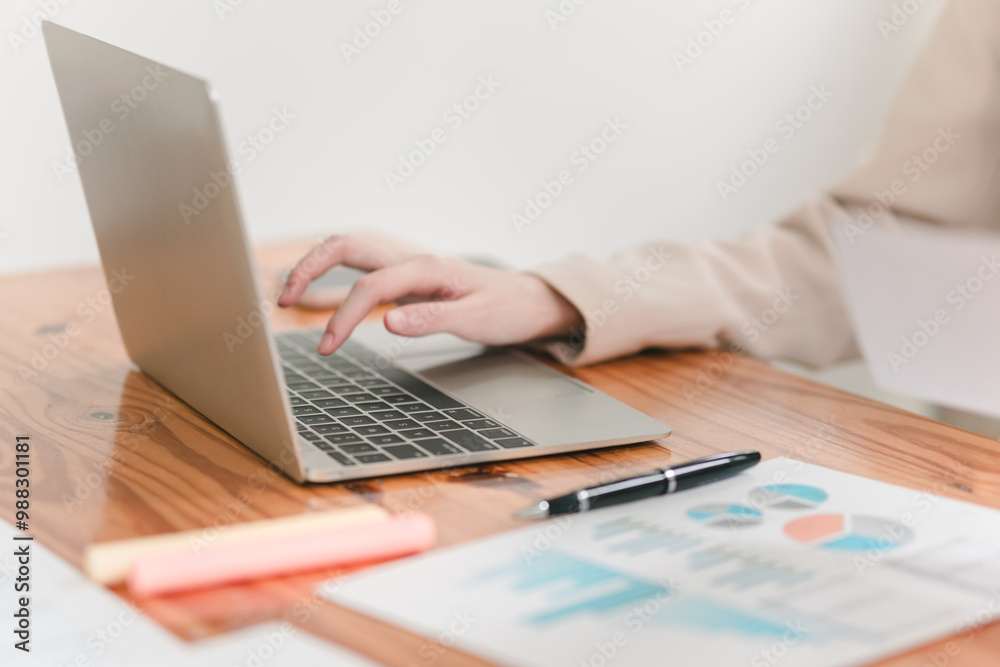 A person uses a laptop on a wooden desk with a pen and colorful charts in view, indicating a work or study scenario.