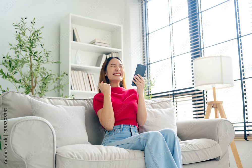 Smiling pretty young Asian woman sitting on cozy couch, using funny mobile apps in living room. Woman at Home, Doing Online Shopping, Messaging Friends, Posting on Social Media Smiling pretty young Asian woman sitting on cozy couch, using funny mobile apps in living room. Woman at Home, Doing Online Shopping, Messaging Friends, Posting on Social Media
