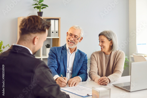 Elderly couple is discussing retirement plans and investment strategies with financial advisor. Elegantly dressed elderly married couple with beaming smiles are signing contract at table in office.