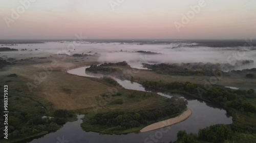 Morning fog begins to creep along the winding river among meadows, fields and forests. In the morning before sunrise.
