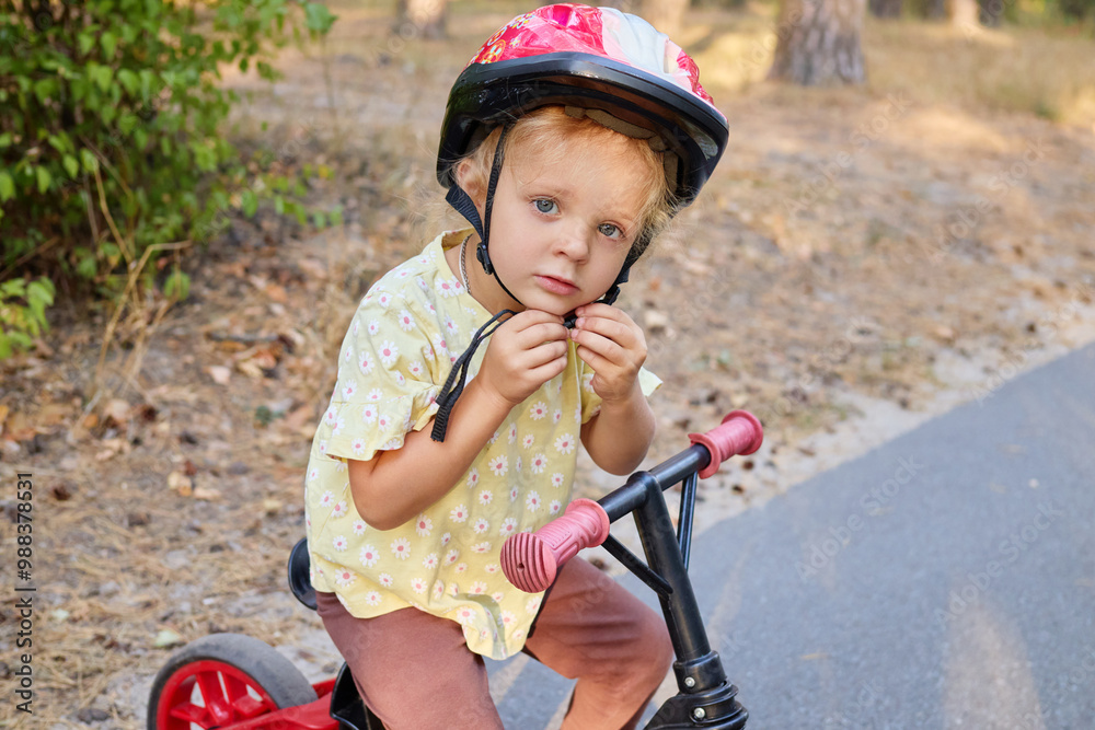 Naklejka premium Little wavy haired girl balancing on her bike along the path in park enjoying the warmth of the sun and the thrill of her first outdoor adventure with push bike