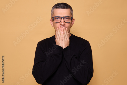 Nervous adult male wearing black jumper and glasses looking at camera with anxiety and frustration has frightened expression feeling scared and confused isolated over beige background