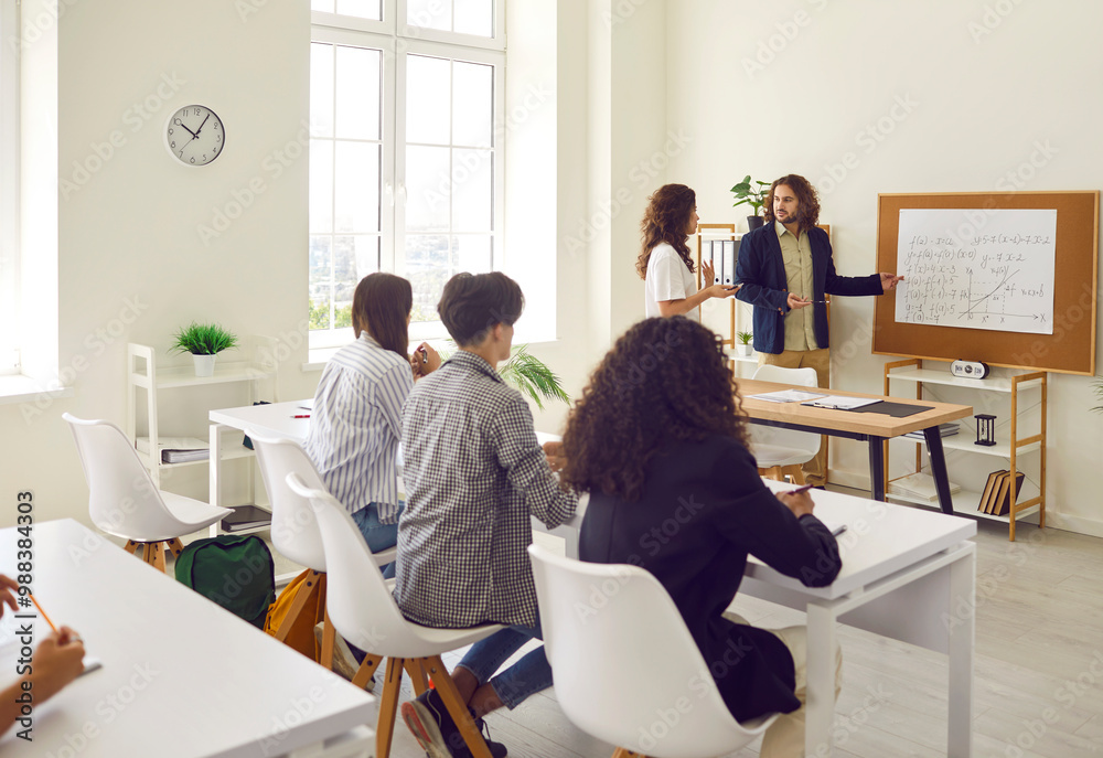 Young girl student standing at the blackboard in classroom and answering the male teacher standing in front of college or high school students during a lesson. Education and learning concept. Young girl student standing at the blackboard in classroom and answering the male teacher standing in front of college or high school students during a lesson. Education and learning concept.