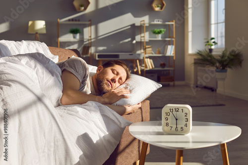 Man is sleeping on the sofa at home with an alarm clock nearby in the morning. The relaxed setting shows him resting before an early wake-up, capturing a peaceful moment in his bedroom.