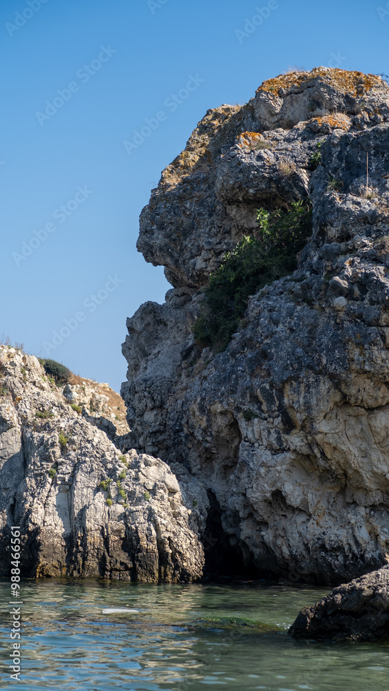 A prominent rocky cliff rises above tranquil waters, surrounded by natural vegetation beneath a bright blue sky at midday A prominent rocky cliff rises above tranquil waters, surrounded by natural vegetation beneath a bright blue sky at midday