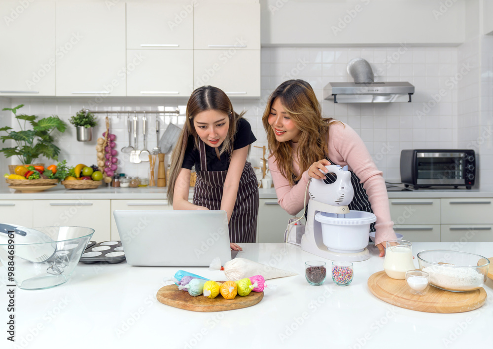 Two young women in an apron make dessert in the kitchen while looking at online recipe guide book on laptop computer.