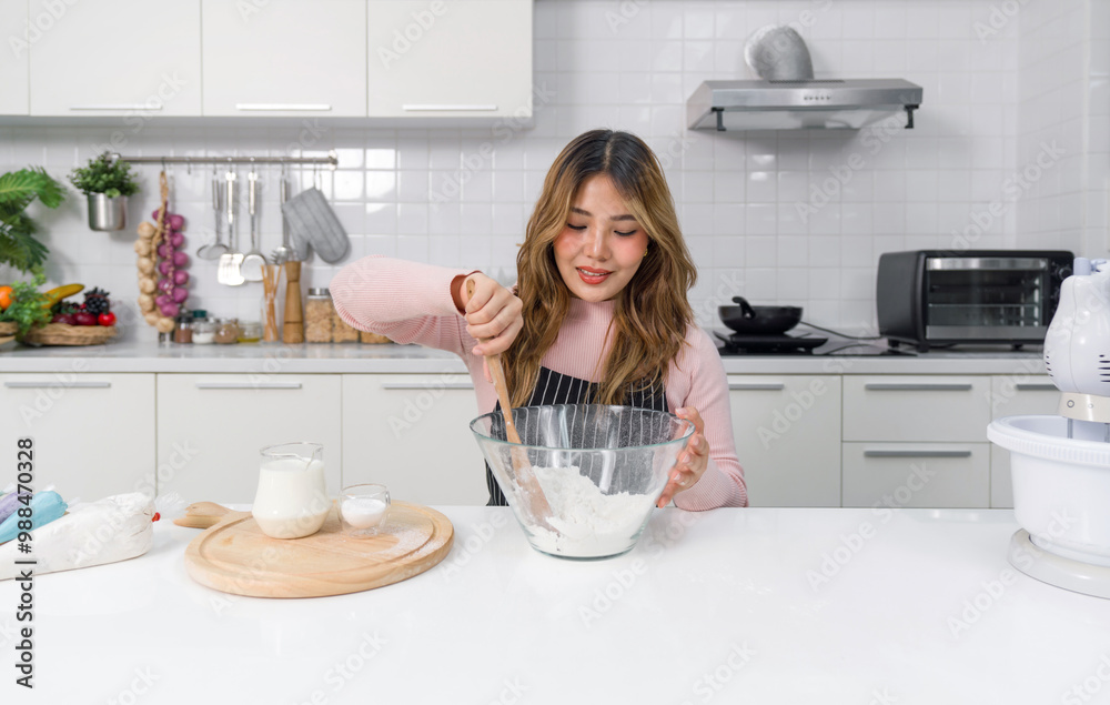 A happy woman in an apron, mixing batter in a clear glass bowl with spatula. Sitting in a clean and modern kitchen. Tray of milk and baking powder placed beside. A happy woman in an apron, mixing batter in a clear glass bowl with spatula. Sitting in a clean and modern kitchen. Tray of milk and baking powder placed beside.