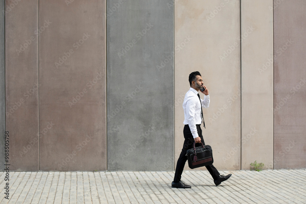 Businessman talking on smart phone and walking through sidewalk near wall Businessman talking on smart phone and walking through sidewalk near wall