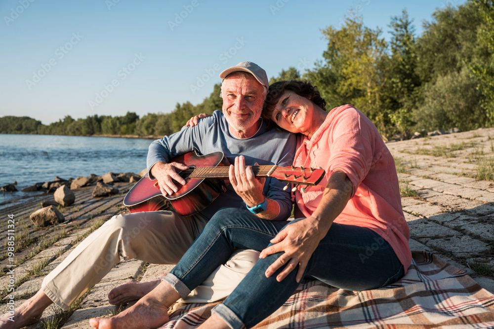 Smiling senior man playing guitar with woman sitting near riverbank on sunny day Smiling senior man playing guitar with woman sitting near riverbank on sunny day