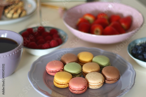 Plate of pastel macarons, cookies and chocolate, cup of tea of coffee, glass of bubble water, various berries, books and accessories on the table. Selective focus, pastel colors.