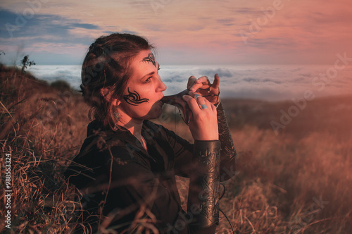 Beautiful girl in gothic style clothes with tattoo on her face plays ocarina in the mountains