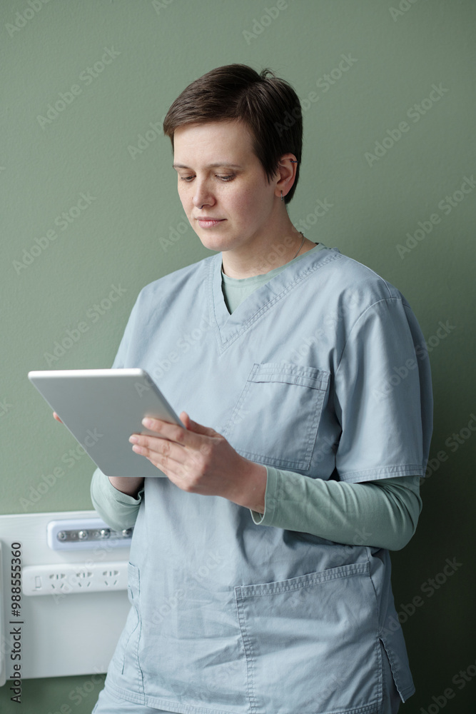 Medical professional in scrubs using a tablet while standing against a green wall reviewing medical data and working in clinical environment Medical professional in scrubs using a tablet while standing against a green wall reviewing medical data and working in clinical environment