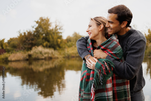 Smiling man hugging girlfriend in blanket near lake in autumn