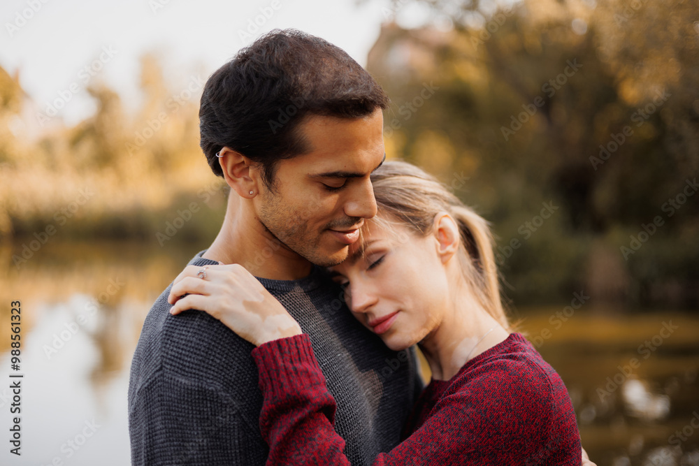 Fototapeta premium Portrait of woman in sweater hugging mixed race boyfriend near blurred lake