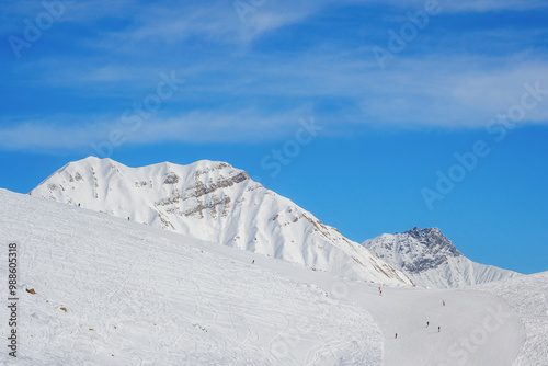 Snowy slopes with ski riders against mountain range in sunny day. Ski resort Gudauri, Georgia. Caucasus Mountains. Aerial view.