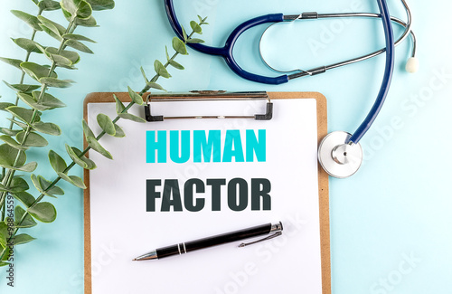 Clipboard with the text human factor on a light blue background, surrounded by a stethoscope, green leaves, and a pen