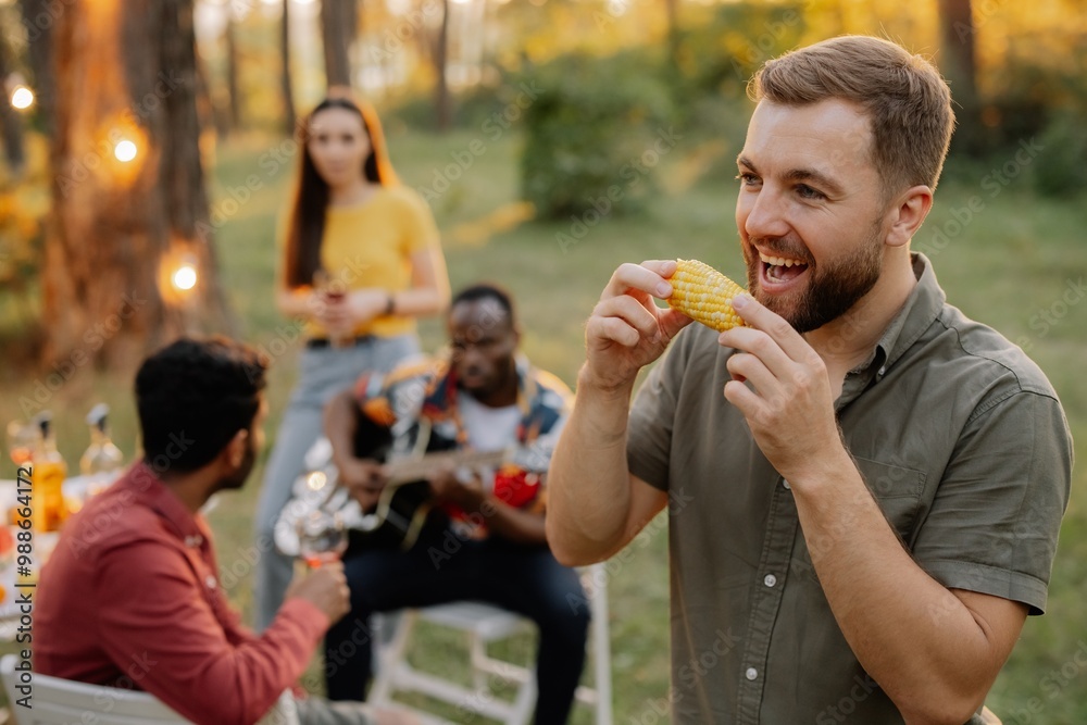 Obraz premium Bearded hipster man eating corn on the background of dinner party of multiracial friends
