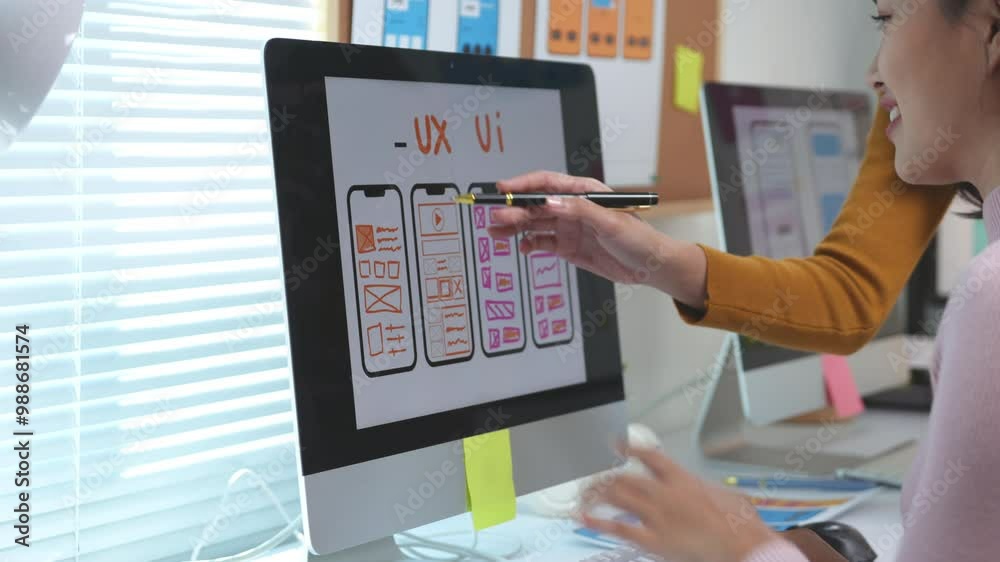 Two female web designers are collaborating on a ux ui design project, using a desktop computer and pointing at the screen