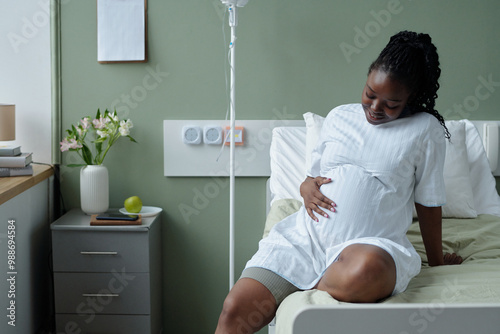 Foto Smiling pregnant woman wearing a white gown sitting on a hospital bed, cradling her stomach and looking down thoughtfully