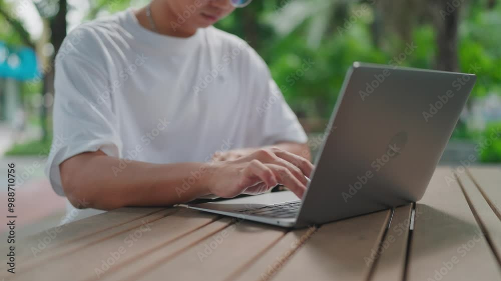 Close up Working on keyboard with laptop, serious looking tan Asian man with glasses studying and taking notes while working outdoors in park with laptop, books and documents on table, daytime, summer Close up Working on keyboard with laptop, serious looking tan Asian man with glasses studying and taking notes while working outdoors in park with laptop, books and documents on table, daytime, summer