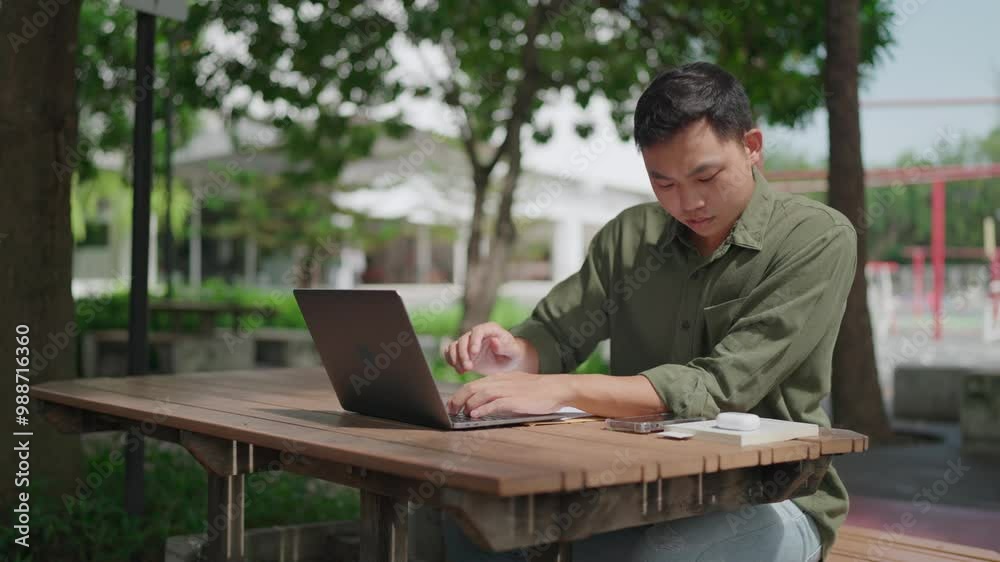 Asian man multitasking outdoors in a park using laptop and smartphone while working on documents on wooden table. Phone has incoming call while working. Freelance, remotely, sitting in city park. Asian man multitasking outdoors in a park using laptop and smartphone while working on documents on wooden table. Phone has incoming call while working. Freelance, remotely, sitting in city park.
