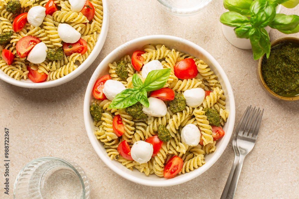 Cold pasta salad with homemade pesto sauce, tomato and mozzarella cheese, decorated with basil leaves on a beige table surface. Directly above. Cold pasta salad with homemade pesto sauce, tomato and mozzarella cheese, decorated with basil leaves on a beige table surface. Directly above.
