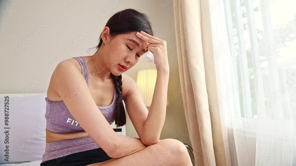 Asian woman in sportswear with cold sitting on bed feeling unwell headache runny nose fever using tissue to wipe her nose. Asian woman in sportswear with cold sitting on bed feeling unwell headache runny nose fever using tissue to wipe her nose.
