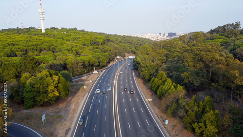Autoestrada em Monsanto, Lisboa, ao pôr do sol com tráfego em movimento. O céu alaranjado ilumina a estrada e os veículos, criando um contraste entre a natureza e a paisagem urbana.