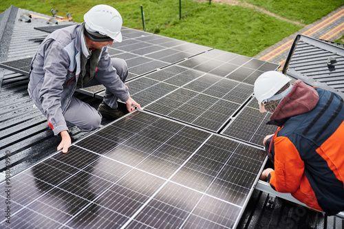 Technicians building photovoltaic solar module station on roof of house. Men engineers in helmets installing solar panel system outdoors. Concept of alternative and renewable energy.