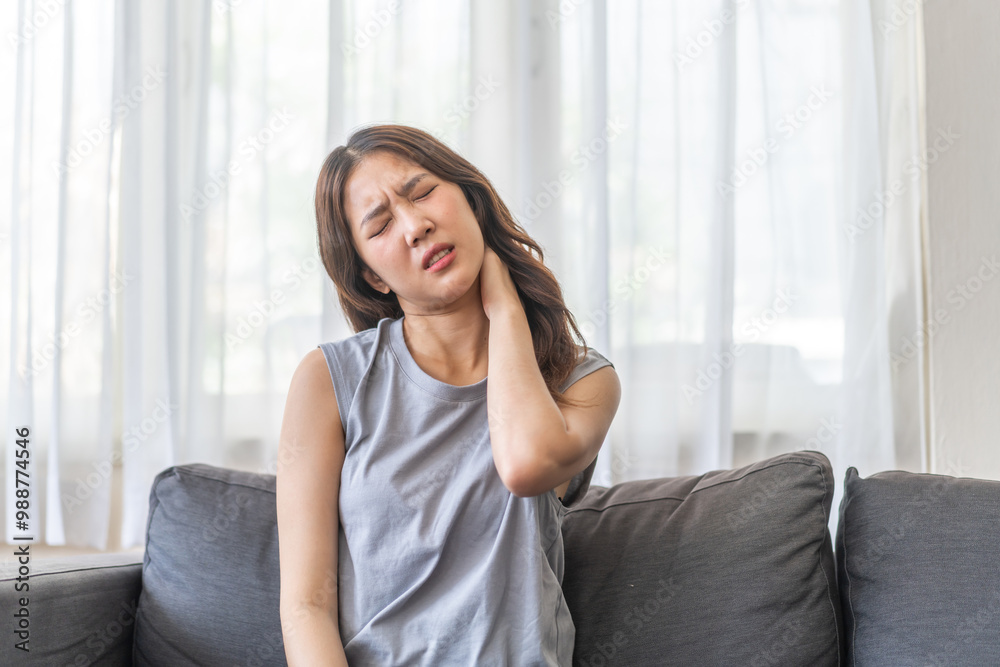 Asian woman sitting on sofa with a painful expression holding her neck, suffering from neck pain indoor home setting, casual clothing, woman health issue, discomfort, stress, fatigue at living room Asian woman sitting on sofa with a painful expression holding her neck, suffering from neck pain indoor home setting, casual clothing, woman health issue, discomfort, stress, fatigue at living room