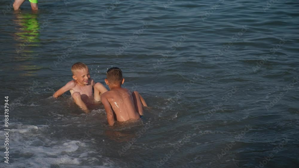 Close-up of two happy boys having fun on the seashore with big waves, they are sitting on the sand and big waves are knocking them down. Children swim in the sea with big waves.