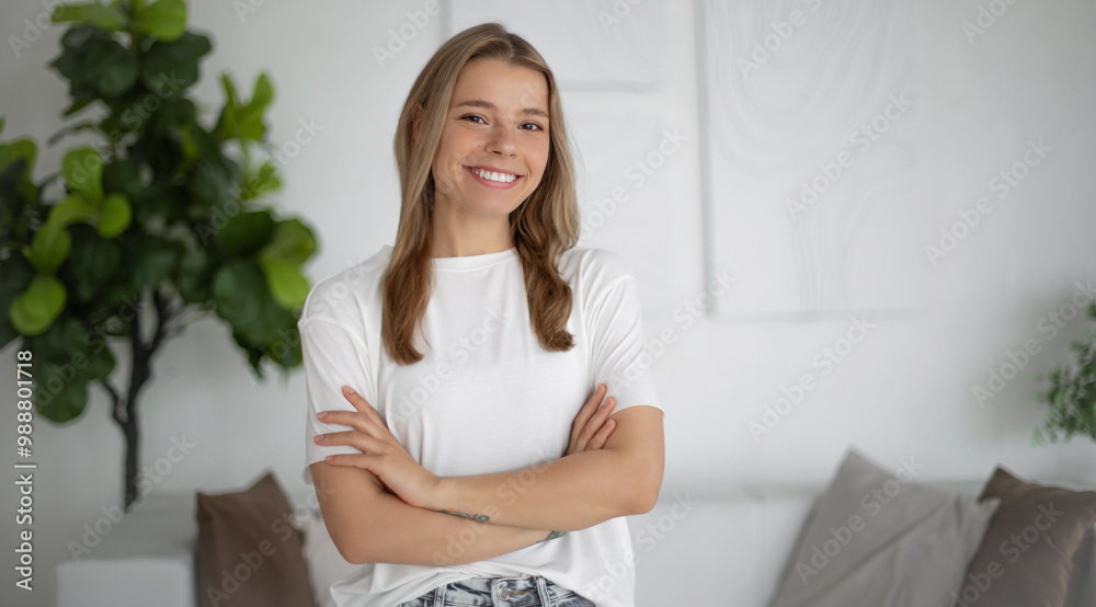 A young woman with long blonde hair is smiling confidently with arms crossed, wearing a white t-shirt. She stands in a modern, bright, and minimalistic living room with plants in the background.