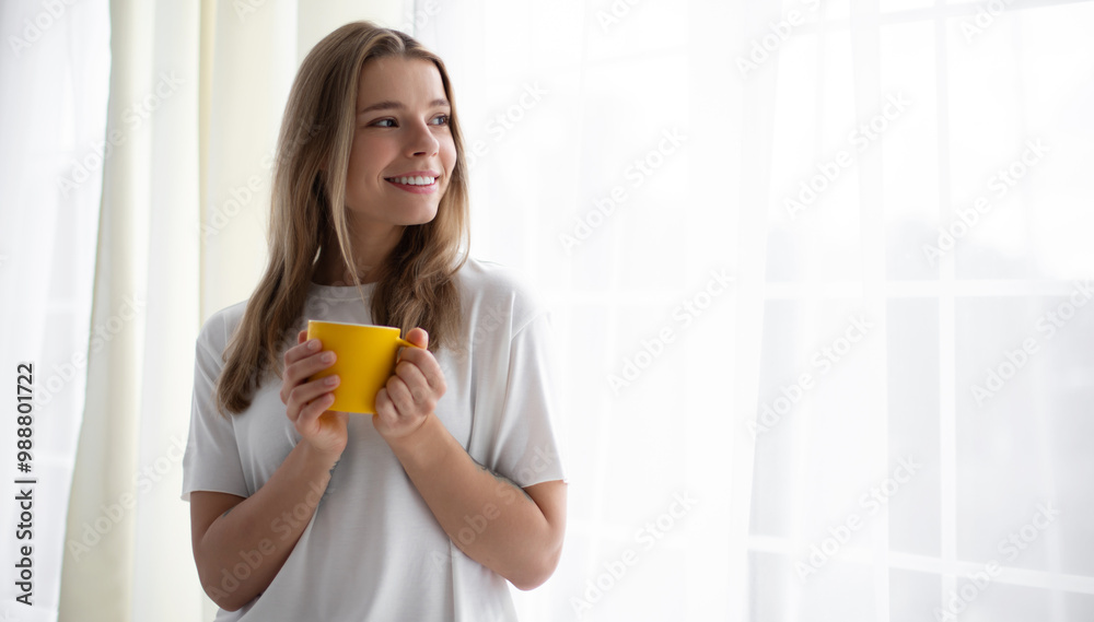 Lovely young woman stands by a bright window, smiling while holding a yellow mug. She wears a white t-shirt, enjoying a peaceful moment in a minimalist, light-filled room, evoking warmth and calm. Lovely young woman stands by a bright window, smiling while holding a yellow mug. She wears a white t-shirt, enjoying a peaceful moment in a minimalist, light-filled room, evoking warmth and calm.