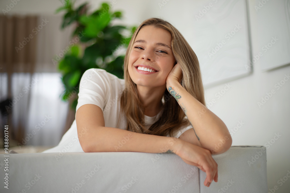 Gorgeous woman with long blonde hair smiles brightly while leaning on couch, wearing white t-shirt. The relaxed, cozy indoor setting features greenery and soft lighting, evoking warmth and happiness Gorgeous woman with long blonde hair smiles brightly while leaning on couch, wearing white t-shirt. The relaxed, cozy indoor setting features greenery and soft lighting, evoking warmth and happiness