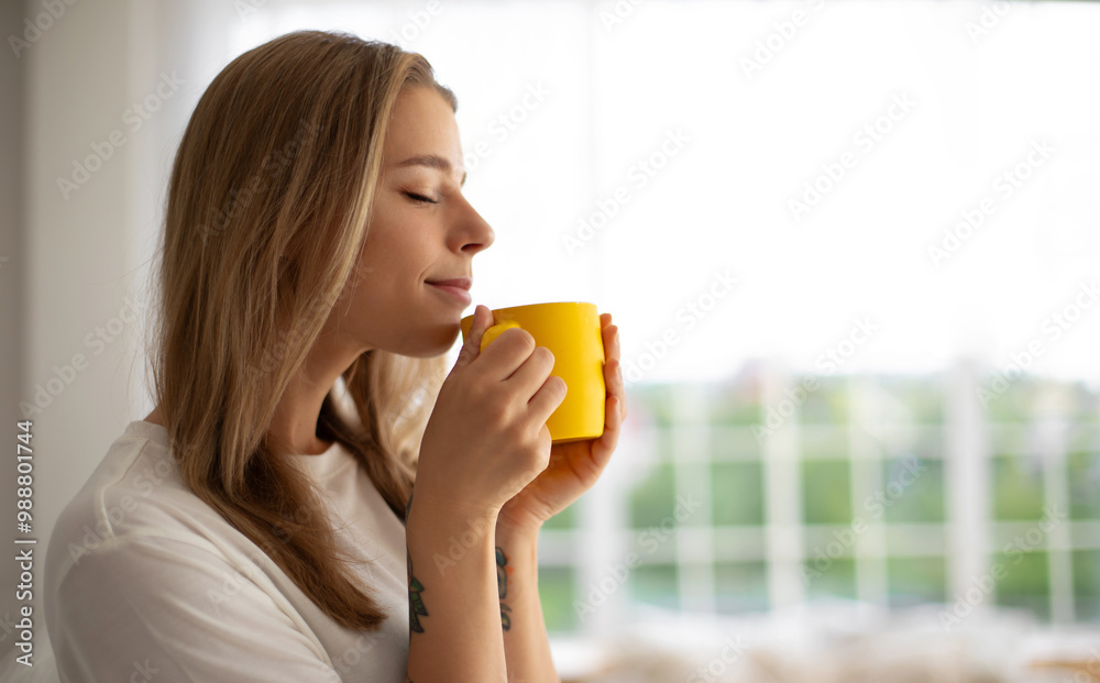 Smiling young woman holds yellow mug, enjoying drink in cozy indoor. She wears a white t-shirt, with soft natural light streaming through large windows in the background, evoking warmth and comfort Smiling young woman holds yellow mug, enjoying drink in cozy indoor. She wears a white t-shirt, with soft natural light streaming through large windows in the background, evoking warmth and comfort