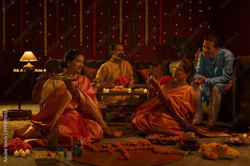 Cheerful senior woman capturing snap of her friend with holding a tray full of diyas during Diwali celebration and their husbands sitting beside and watching