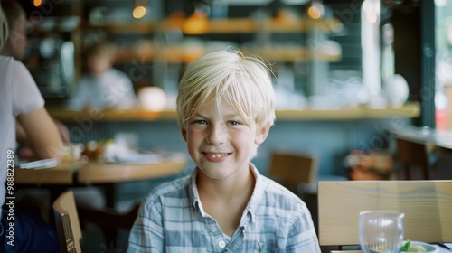A young boy with blonde hair smiles sweetly at the camera while sitting in a cozy cafe, creating a warm and friendly atmosphere.