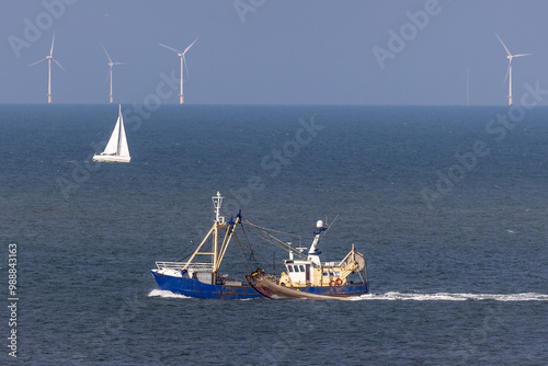 Fishing vessel with lifted dragnets navigates waters near offshore wind turbines with a yacht visible in the distance during a sunny day photo