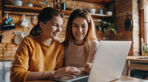 Two young women share a laugh while working on a laptop in a cozy kitchen, illustrating a moment of friendship and collaboration in a casual setting.