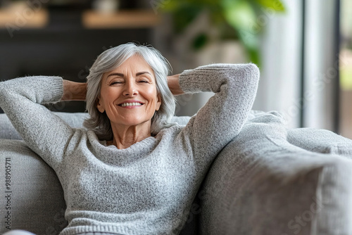 Cinematic medium shot of an older woman sitting on the couch with her hands behind her head, smiling joyfully as she relaxes at home.