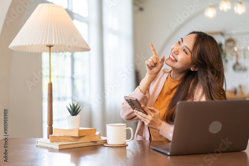 Cheerful asian woman pointing up holding her smartphone and smiling, working remotely at home with a laptop, casual lifestyle, drinking coffee, enjoying work from home environment, positive energy photo