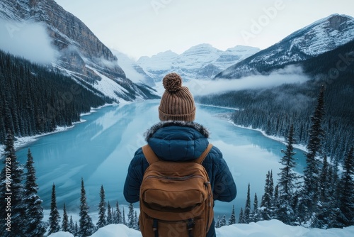 An individual wearing a beanie and backpack stands atop a viewpoint, gazing at the stunning scene of a frozen lake surrounded by snow-covered mountains with towering pine trees.